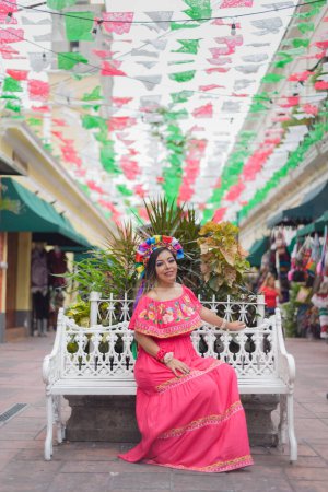 Mexikanerin sitzt in traditioneller Kleidung. Straße dekoriert mit den Farben der mexikanischen Flagge. Feier zum Cinco de Mayo.