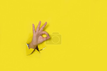 Foto de Mano haciendo una señal de OK, saliendo del agujero en un fondo de papel amarillo roto. - Imagen libre de derechos