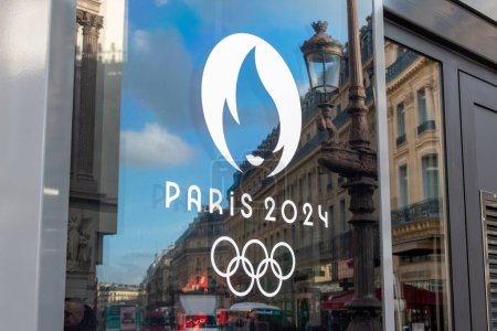 Téléchargez les photos : Paris, France - 6 décembre 2023 : Gros plan sur le logo Paris 2024, symbole des Jeux Olympiques d'été de 2024 à Paris, sur une fenêtre avec le reflet d'une rue parisienne - en image libre de droit