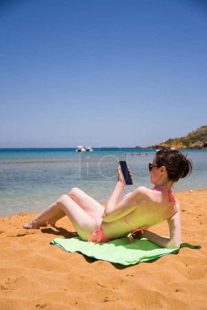 Foto de Gozo, Malta - 19 junio, 2023: Niña con tez muy clara tomando el sol con smartphone en la mano - Imagen libre de derechos