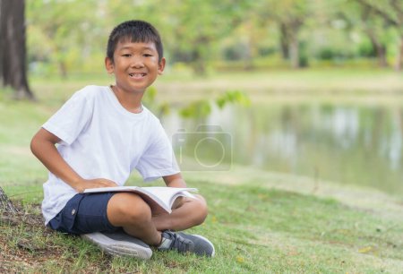 Téléchargez les photos : Mignon asiatique garçon est heureux il assis et lecture sur herbe sol dans jardin avec flou feuilles arrière-plan. - en image libre de droit