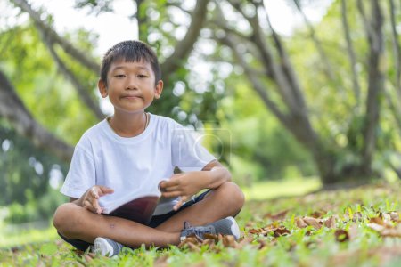 Téléchargez les photos : Mignon asiatique garçon est heureux il assis et lecture sur herbe sol dans jardin avec flou feuilles arrière-plan. - en image libre de droit