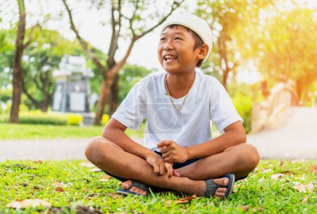 Téléchargez les photos : Garçon mignon en T-shirt blanc est heureux, il est assis sur le sol d'herbe au parc de la ville le matin avec des feuilles vertes floues et de l'herbe pour le fond. - en image libre de droit