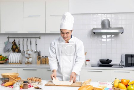 Téléchargez les photos : Portrait de chef masculin avec chapeau et tablier pétrissant la pâte. tout en préparant dans la cuisine à la maison. Chef asiatique en tabliers blancs faire de la pâtisserie sur la table dans la cuisine heureusement. - en image libre de droit