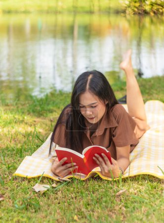 Photo for Attractive woman lying down and reading red book on yellow mat at city park with copy space. Enjoying free time concept. - Royalty Free Image