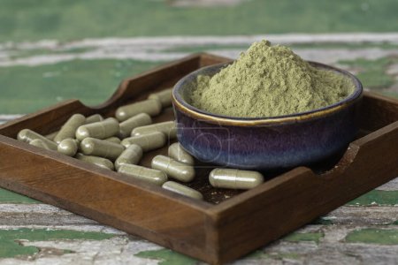 Photo for Natural plant herbal extract powder in ceramic bowl and herbal capsules in wooden saucer and on old green wooden background. - Royalty Free Image