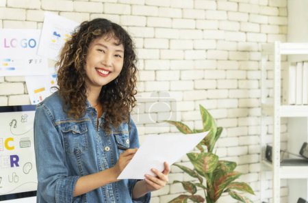 Photo for Portrait of attractive female graphic designer in denim shirt. She is holding piece of paper and smiling happily. - Royalty Free Image