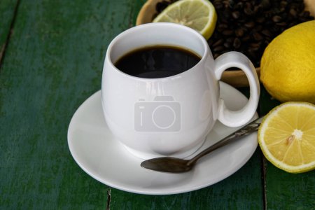 Foto de Café negro caliente en taza blanca, con limón amarillo, granos de café en platillo de madera y rodajas de limón sobre fondo de madera verde viejo. - Imagen libre de derechos