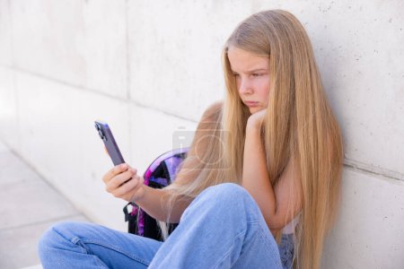Téléchargez les photos : Adolescente assise seule dehors et regardant le téléphone mobile - en image libre de droit