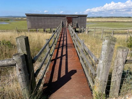 A bird hide on a marsh nature reserve.A walkway leads up to the wooden oblong building with fields and water in background.j
