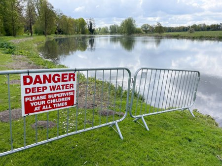 Fencing and crowd control barriers has been placed by a lake to prevent access and a sign says 'Danger Deep Water.Please supervise your children at all times'.