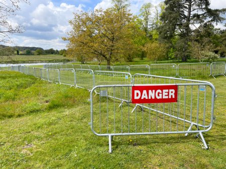 Un panneau indique "Danger" et des barrières de contrôle de foule en métal interconnectés marquent une zone d'accès dans un champ d'herbe avec des arbres et des campagnes en arrière-plan
