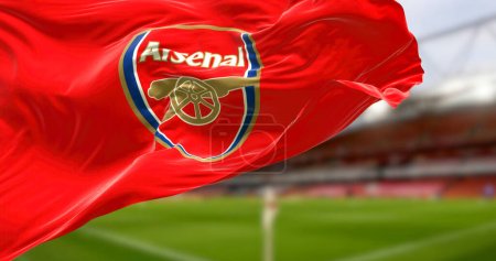 Foto de London, UK, DEC 2022: flag of Arsenal Football Club waving with the Emirates Stadium blurred in the background. Rippled Fabric. Textured background. Selective focus. Realistic 3d illustration - Imagen libre de derechos