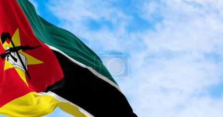 Nahaufnahme der mosambikanischen Nationalflagge, die an einem klaren Tag geschwenkt wird. Grüne, schwarze und gelbe Streifen, rotes Dreieck, gelber Stern, gekreuztes Buch, Hacke und AK-47. 3D Illustration rendern. Selektiver Fokus. Nahaufnahme