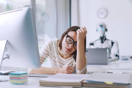 Foto de Decepcionada mujer de negocios estresada y robot AI sentado en su escritorio en la oficina, la mujer está preocupada y pensativa - Imagen libre de derechos