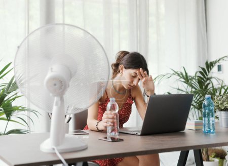 Téléchargez les photos : Femme assise au bureau à la maison lors d'une vague de chaleur estivale, elle se refroidit avec un ventilateur électrique - en image libre de droit
