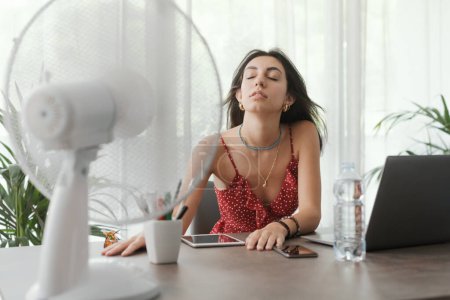 Téléchargez les photos : Femme assise au bureau à la maison et travaillant avec un ordinateur, elle se refroidit avec un ventilateur électrique - en image libre de droit