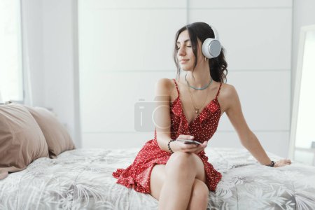 Photo for Young woman wearing headphones and sitting on the bed, she is listening to music online - Royalty Free Image