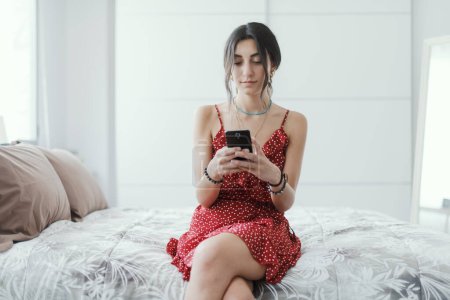 Photo for Young fashionable woman sitting on the bed at home and connecting online with her smartphone, she is chatting and smiling - Royalty Free Image