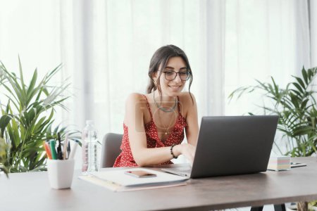 Téléchargez les photos : Jeune étudiante assise au bureau et connectée à son ordinateur portable, apprentissage en ligne et concept de médias sociaux - en image libre de droit