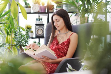 Foto de Mujer atractiva joven relajándose en casa rodeada de hermosas plantas, ella está sentada y leyendo un libro - Imagen libre de derechos