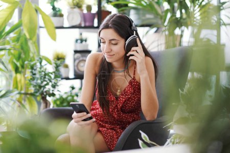 Foto de Mujer joven que se relaja en casa rodeada de plantas exuberantes, lleva auriculares y se conecta con su teléfono inteligente - Imagen libre de derechos
