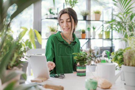 Téléchargez les photos : Femme prenant soin de ses plantes à la maison, elle regarde des vidéos de jardinage en ligne sur sa tablette numérique - en image libre de droit