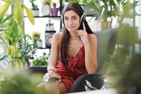 Foto de Mujer joven que se relaja en casa rodeada de plantas exuberantes, lleva auriculares y se conecta con su teléfono inteligente - Imagen libre de derechos