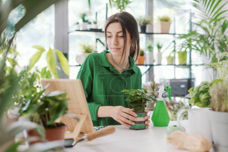 Téléchargez les photos : Femme prenant soin de ses plantes à la maison, elle regarde des vidéos de jardinage en ligne sur sa tablette numérique - en image libre de droit