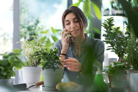 Téléchargez les photos : Femme relaxante entourée de plantes à la maison, elle reçoit un appel téléphonique avec son smartphone - en image libre de droit