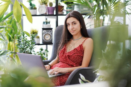 Foto de Mujer atractiva joven relajándose en casa rodeada de hermosas plantas, ella está trabajando con su computadora portátil - Imagen libre de derechos