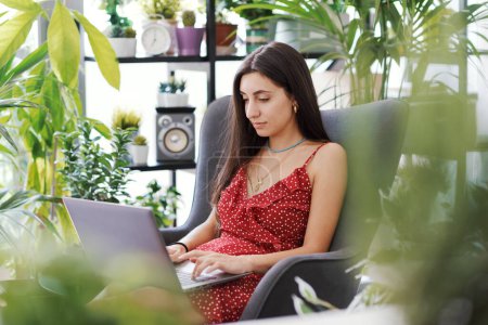 Photo for Young attractive woman relaxing at home surrounded by beautiful plants, she is working with her laptop - Royalty Free Image
