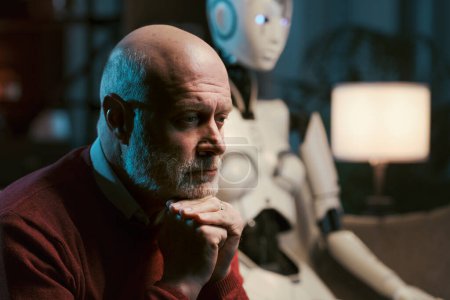 Photo for Sad lonely man at home alone sitting on the couch with his AI robot - Royalty Free Image