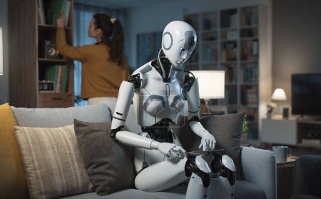 Foto de Una chica joven y bonita y su robot equipado con inteligencia artificial pasan una noche tranquila juntos, leyendo y conversando. Concepto de utilidad de la IA para servir a los humanos. - Imagen libre de derechos