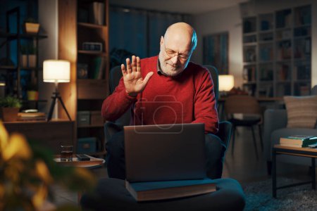 Foto de Hombre mayor sentado en el sillón en casa y usando su computadora portátil, está teniendo una videollamada y saludando - Imagen libre de derechos