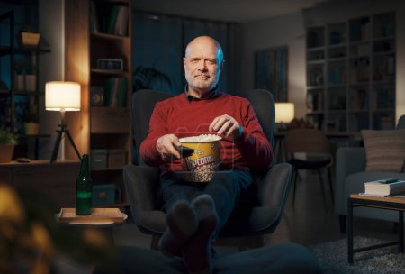 Téléchargez les photos : Heureux homme âgé se détendre à la maison et regarder la télévision, il mange du pop-corn et souriant - en image libre de droit