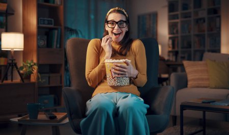 Téléchargez les photos : Femme assise sur le fauteuil à la maison et regardant un film comique, elle mange du pop-corn et sourit - en image libre de droit