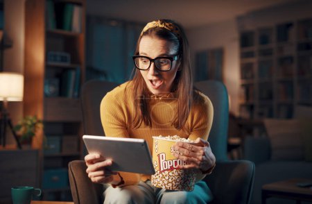 Téléchargez les photos : Femme heureuse assise dans le salon et se connectant à sa tablette numérique, elle regarde des films en ligne - en image libre de droit