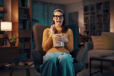 Photo for Woman sitting on the armchair at home and watching a horror movie, she is eating popcorn and feeling shocked - Royalty Free Image