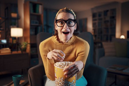 Photo for Scared woman watching a horror movie on TV and eating popcorn - Royalty Free Image