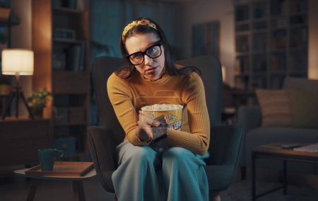 Téléchargez les photos : Femme triste ennuyée assise dans le salon et regardant la télévision, elle tient la télécommande et regarde la caméra - en image libre de droit
