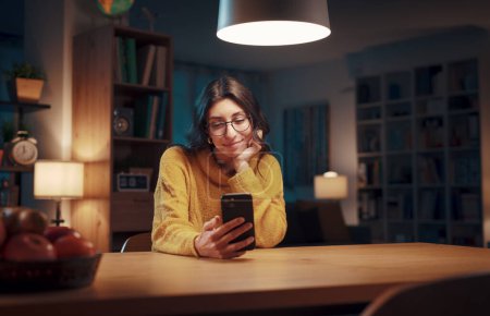 Foto de Joven mujer feliz sentada en la sala de estar por la noche y charlando con su teléfono inteligente, estilo de vida y concepto de tecnología - Imagen libre de derechos