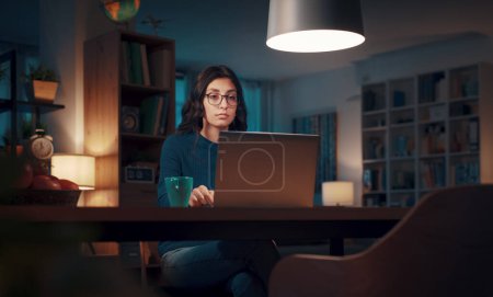 Foto de Mujer joven sentada en su escritorio en casa y trabajando con un ordenador portátil tarde en la noche, el trabajo desde el concepto de casa - Imagen libre de derechos