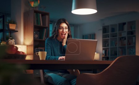Foto de Mujer joven y feliz sentada en su escritorio y las redes sociales con su portátil por la noche - Imagen libre de derechos