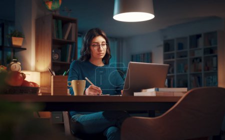 Foto de Mujer sentada en su escritorio y conectada con su portátil a altas horas de la noche, está estudiando y asistiendo a clases en línea - Imagen libre de derechos