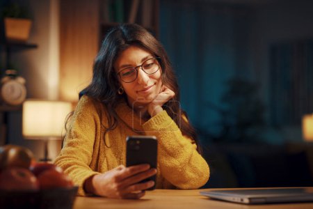 Téléchargez les photos : Jeune femme heureuse assise dans le salon la nuit et bavarder avec son smartphone, style de vie et concept de technologie - en image libre de droit