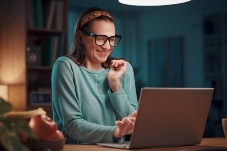 Téléchargez les photos : Heureuse femme excitée assise à son bureau à la maison et se connectant avec un ordinateur portable : elle est en ligne de réseautage social - en image libre de droit
