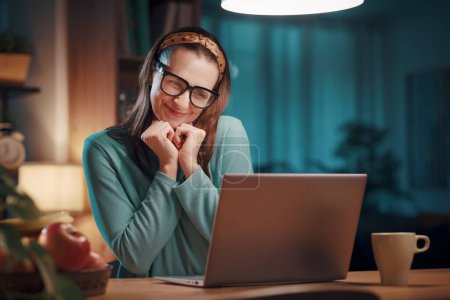 Foto de Mujer romántica feliz sentada en su escritorio en casa y conectándose con un ordenador portátil, ella está sonriendo y mirando a la pantalla de la computadora - Imagen libre de derechos