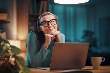 Photo for Happy romantic woman sitting at her desk at home and connecting with a laptop, she is smiling and looking away - Royalty Free Image