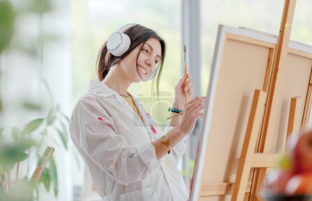 Foto de Mujer joven y feliz escuchando música con auriculares y pintando sobre lienzo en el estudio de arte - Imagen libre de derechos